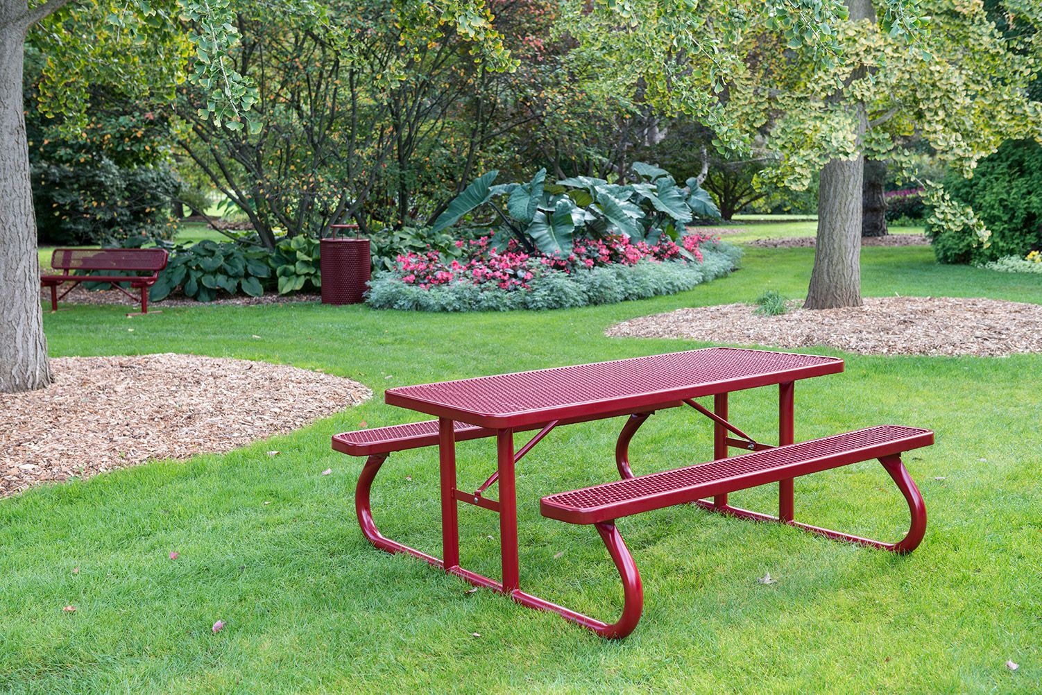 A red picnic table sits in a grassy area amongst trees with tan mulch around them and plants. A matching red bench and trashcan are also visible.